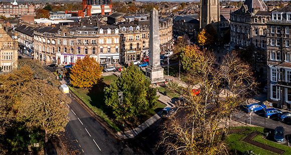 aerial view of a town square featuring a tall monument surrounded by buildings trees and vehicles autumn scenery on a sunny day the square includes six trees and is a popular gathering spot